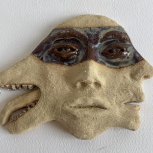 Può includere: Una scultura da parete in ceramica beige con un design a più volti. Il volto centrale presenta una zona oculare liscia smaltata in marrone scuro. A sinistra, emerge un secondo profilo con denti e una lingua visibili.