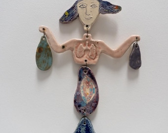 May include: A ceramic mobile sculpture of a stylized female figure with blue hair, pink torso, and blue and purple lower body. The figure is holding two small, teardrop-shaped ceramic pieces in each hand.