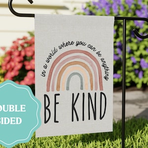 May include: A double-sided garden flag with the text "BE KIND" in black block letters. Above the text is a watercolor rainbow. The flag also has the text "in a world where you can be anything". The flag is hanging on a black metal stand.