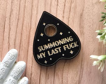 Puede incluir: Plancheta negra con el texto "SUMMONING MY LAST FUCK" en color claro. La plancheta tiene un agujero y recortes en forma de estrella. El fondo es una superficie de madera marrón claro.