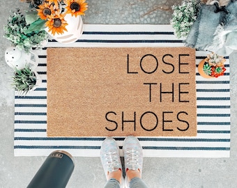 Könnte beinhalten: Eine rechteckige, hellbraune Kokosfasermatte mit dem schwarzen Schriftzug LOSE THE SHOES, die auf einer weiß-dunkelblau gestreiften Matte liegt. Die Fußmatte befindet sich auf einem Betonboden mit verschiedenen Pflanzen und einem dunkelgrauen Becher.