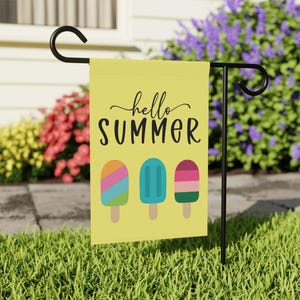 May include: A yellow garden flag hanging on a black metal stand. The flag features the text "hello SUMMER" in black script and block lettering above three colorful popsicle illustrations with stripes in blue, pink, orange, green, and red.