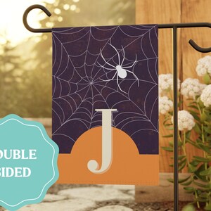 May include: A rectangular garden flag featuring a dark purple background with a white spider web pattern and a white spider. The lower portion has an orange semi-circle and a large white letter J. The flag is displayed on a black metal stand.