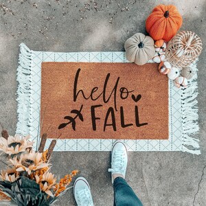 May include: A brown doormat with the words "hello FALL" in black script, a leaf design, and a white and gray patterned border. Decorative pumpkins in various sizes and textures are arranged on the mat, with a bouquet of sunflowers in the corner.