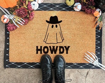 Op de afbeelding: Een rechthoekige deurmat met een zwart-witte rand en een bruin centrum. De mat heeft een spook met een cowboyhoed en het woord "HOWDY" in het zwart. Zwarte laarzen en panty's met patronen zijn zichtbaar.