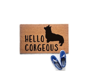 May include: A rectangular tan coir doormat featuring a black silhouette of a Corgi dog and the black text "HELLO CORGEOUS" printed on the surface. A pair of blue canvas boat shoes with white laces are positioned near the bottom right corner.