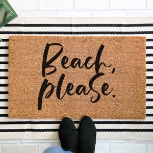 May include: A rectangular doormat with the words "Beach, please." in black script. The mat is a light brown colour and sits on a black and white striped rug. Black shoes are visible at the bottom of the image.