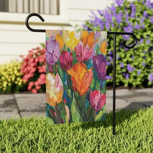 May include: A rectangular garden flag featuring an impressionistic painting of vibrant tulips in shades of orange, yellow, pink, purple, and white, displayed on a black metal stand with a curved top hook, set against a green lawn.
