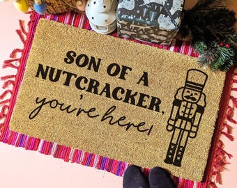 May include: A rectangular tan coir doormat featuring the black printed text SON OF A NUTCRACKER, you're here! and a black graphic illustration of a nutcracker soldier standing on the right side of the mat, layered over a red and pink fringed rug.