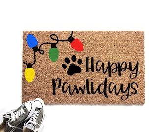 May include: A rectangular tan coir doormat featuring a black paw print and the text Happy Pawlidays in black script. A string of colorful Christmas lights in blue, green, red, and yellow is printed above the text. A pair of gray sneakers is visible.