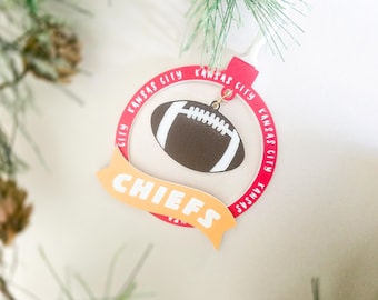 May include: A circular Kansas City Chiefs football ornament featuring a red border with the text Kansas City repeated around the edge. A brown football with white laces hangs in the center, and a yellow banner with the word CHIEFS is attached.
