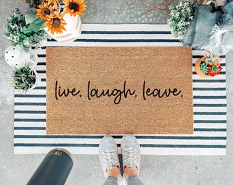 May include: A rectangular tan coir doormat featuring the black text "live. laugh. leave." in a cursive script font. The doormat is layered over a larger white and navy blue striped rectangular rug, creating a layered entryway floor decor set.