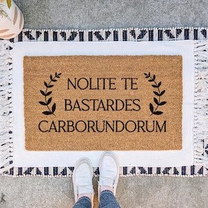 May include: A rectangular coir doormat with the phrase "Nolite Te Bastardes Carborundorum" in black script, framed by laurel branches. The mat is set on a white and patterned rug with fringe, with a pair of white shoes visible.