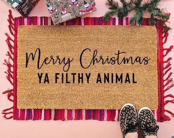 May include: A rectangular tan coir doormat featuring the black text Merry Christmas YA FILTHY ANIMAL. The mat is layered over a red, pink, and purple fringed rug. The design includes a festive holiday theme with gift wrapping and greenery.