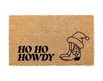 May include: A rectangular tan coir doormat featuring the black text HO HO HOWDY on the left side and a black line art illustration of a cowboy boot wearing a Santa hat on the right side. The mat has a coarse, natural fiber texture throughout.