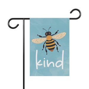 May include: A rectangular garden flag hanging on a black metal stand. The flag features a light blue background with a textured effect, a central illustration of a bee with black and yellow stripes, and the white lowercase text "kind" below.