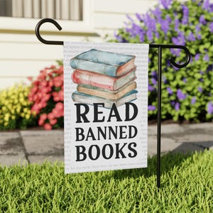 May include: A white garden flag with the words "READ BANNED BOOKS" in bold black letters. The flag features a watercolor illustration of a stack of books in shades of blue, pink, and tan. The flag is displayed on a black metal stand.