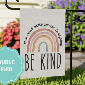 May include: A rectangular garden flag featuring a muted rainbow graphic in shades of pink, tan, and blue. The text reads, "in a world where you can be anything BE KIND" in black lettering. A teal scalloped badge states "DOUBLE SIDED" in white.