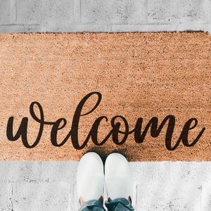 May include: A rectangular doormat made of natural fibres with the word "welcome" in a dark brown script. The mat is placed on a white brick surface, with a person's feet wearing white shoes visible.