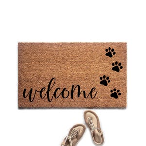 May include: A rectangular brown doormat with the word "welcome" in black script. Black paw prints are on the right side. Two pairs of beige sandals are at the bottom.