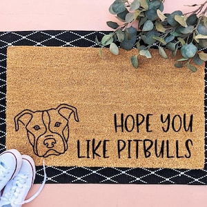 Peut inclure: Paillasson rectangulaire avec un centre beige et une bordure noire. Le tapis présente un dessin au trait d'un visage de pitbull et le texte "HOPE YOU LIKE PITBULLS" en noir. Des baskets blanches sont dans le coin inférieur gauche.