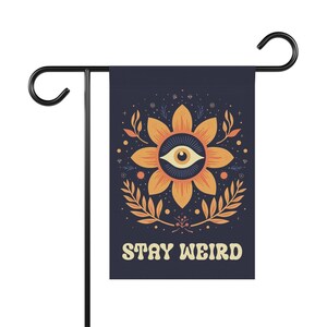 May include: A navy blue garden flag featuring a floral design with an eye in the center, surrounded by orange petals and leaves. The text "STAY WEIRD" is in a retro font. The flag is mounted on a black metal stand.