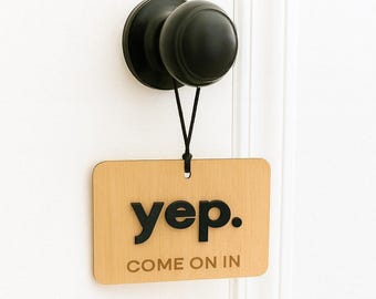 May include: A rectangular light wood sign hanging from a matte black round door knob. The sign features the black text "yep." in a bold sans-serif font above the smaller, thinner text "COME ON IN" in a matching black color.