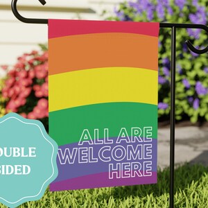 May include: A rectangular garden flag featuring horizontal stripes in red, orange, yellow, green, blue, and purple. The text ALL ARE WELCOME HERE is printed in white capital letters on the lower half. A teal scalloped badge reads DOUBLE SIDED.