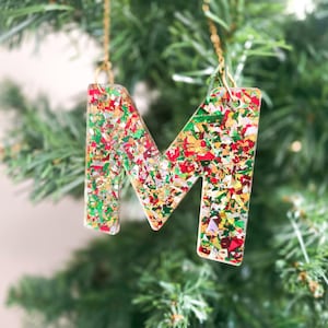 Close-up of a chunky glitter resin letter M ornament with red, green, and gold flakes, hanging from a gold chain on a Christmas tree.