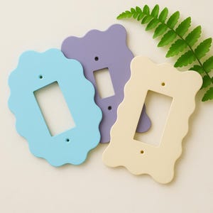 May include: Three decorative light switch covers in pastel colors: light blue, lavender, and cream. Each cover has a scalloped edge and a rectangular opening for the switch. A green fern leaf is in the background.