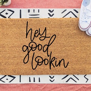May include: A rectangular doormat made of natural coir fibre, with the phrase "hey good lookin" in black cursive script. The mat is placed on a patterned rug with white trainers on top.