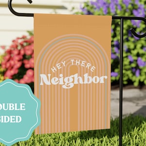 May include: A rectangular garden flag in a muted orange hue featuring a series of concentric arches in lighter shades. The text "HEY THERE" is printed in white above the word "Neighbor" in a large, bold, white, rounded serif font.