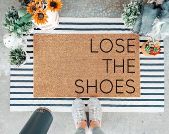 Puede incluir: Una alfombra rectangular con las palabras "LOSE THE SHOES" en letras negras. La alfombra está hecha de un material de fibra natural y se coloca sobre una alfombra a rayas. La imagen también incluye un par de zapatillas y plantas decorativas.