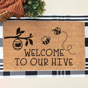 May include: A rectangular doormat with a brown coir surface features a bee-themed design. The design includes a branch with a beehive, two bees, and the text "WELCOME TO OUR HIVE" in black. The mat is set on a black and white checkered rug.