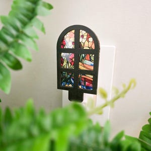 May include: A plug-in night light featuring a black arched frame with a grid design. The six window panes display a colorful stained glass pattern of red, yellow, and blue flowers with green stems and leaves set against a light background.