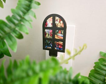 May include: A plug-in night light featuring a black arched frame with a grid design. The six window panes display a colorful stained glass pattern of red, yellow, and blue flowers with green stems and leaves set against a light background.