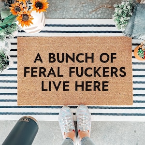 May include: A rectangular tan coir doormat with black text that reads A BUNCH OF FERAL FUCKERS LIVE HERE. The mat is layered over a white and navy blue striped rug. The text is centred in a bold, sans-serif font across three distinct lines.