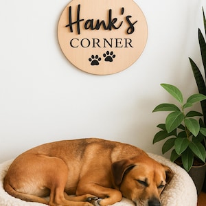 Könnte beinhalten: Ein rundes Wand-Schild aus hellem Holz mit dem schwarzen Schriftzug "Hank's CORNER" in einer Kombination aus Schreib- und Druckschrift, ergänzt durch zwei schwarze Pfotenabdrücke. Das Schild hängt an einer Wand über einem schlafenden beigen Hund.