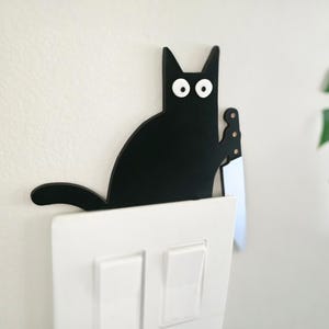 May include: A black cat-shaped light switch cover with white eyes and a knife. The cat is perched above a white light switch. The background is a plain white wall with a green plant in the corner.