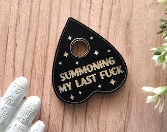 May include: A black heart-shaped planchette featuring a circular cutout at the top and engraved gold-colored text that reads SUMMONING MY LAST FUCK. The surface is decorated with small four-pointed star icons scattered around the lettering.