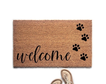 May include: A rectangular brown coir doormat featuring the word welcome in black cursive script and four black paw prints arranged in a diagonal line on the right side, accompanied by a pair of light-colored thong sandals positioned below.