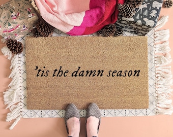 May include: A rectangular tan coir doormat featuring the black text 'tis the damn season centered on the surface. The mat is placed on a white woven rug with fringe edges, surrounded by pinecones and gift-wrapped boxes with holiday patterns.