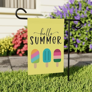 May include: Yellow garden flag with the words "hello SUMMER" and three colorful popsicle illustrations. The flag is hanging on a black metal stand in a grassy yard with flowers in the background.