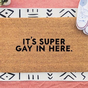 May include: A rectangular doormat with the text "IT'S SUPER GAY IN HERE" in black. The mat is brown and sits on a patterned rug with white and black geometric designs. White sneakers with red trim are on the right.