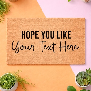 a textured door mat with the phrase "HOPE YOU LIKE Your Text Here" written on it, surrounded by various potted plants and a pink background.