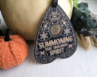 May include: A black planchette-shaped ornament featuring gold engraved details, including an eye, snowflakes, and the text "SUMMONING THE HOLIDAY SPIRIT." The design includes decorative swirls and a central circular cutout, suspended by a black cord.