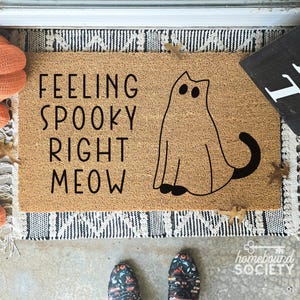Peut inclure: Un paillasson rectangulaire avec le texte "FEELING SPOOKY RIGHT MEOW" et un contour de chat noir. Le tapis est sur un tapis à motifs avec des citrouilles oranges et des feuilles d'automne. Un panneau noir est visible.