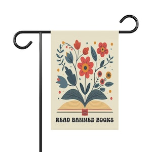 May include: A garden flag with a floral design emerging from an open book. The flowers are red, orange, and yellow with blue-green leaves. The flag has the text "READ BANNED BOOKS" in a retro font. The flag is displayed on a black metal stand.