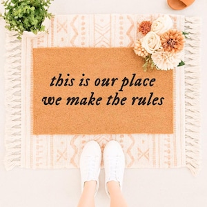 May include: A rectangular brown coir doormat featuring the black text 'this is our place we make the rules' centered on the surface. The mat is layered over a cream-colored woven rug with a geometric pattern and fringed edges on both sides.