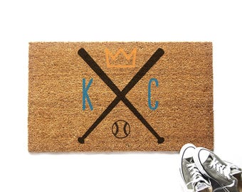 Könnte beinhalten: Eine rechteckige Fußmatte aus Kokosfaser mit Baseball-Motiv. Die Matte zeigt gekreuzte Schläger, einen Baseball und die Buchstaben "KC" in Blau, mit einer goldenen Krone darüber. Ein Paar graue Converse-Sneaker befindet sich in der unteren rechten Ecke.